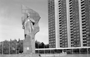 A large statue of a man stands prominently in an open plaza, surrounded by low barricades with a banner.