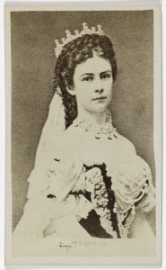 A historical sepia-toned portrait of a woman wearing an elaborate dress and a crown with a lace veil. She gazes directly at the camera.